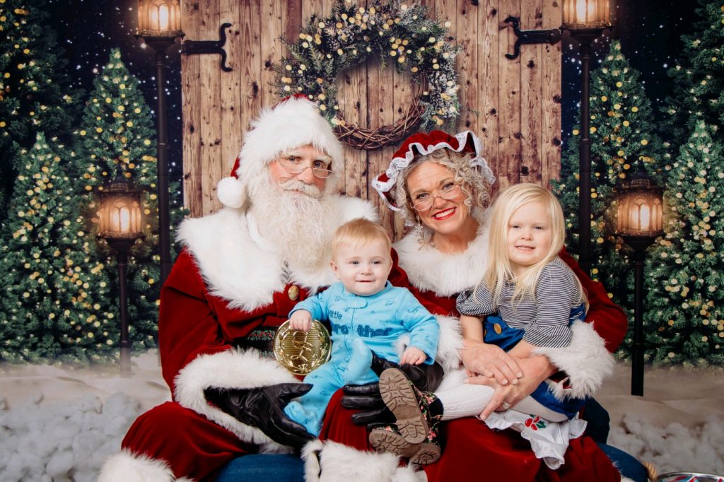 One baby boy in blue, one blonde haired girl in blue sitting on Santa and Mrs. Claus' lap in front of Christmas trees and wooden door with wreath. 