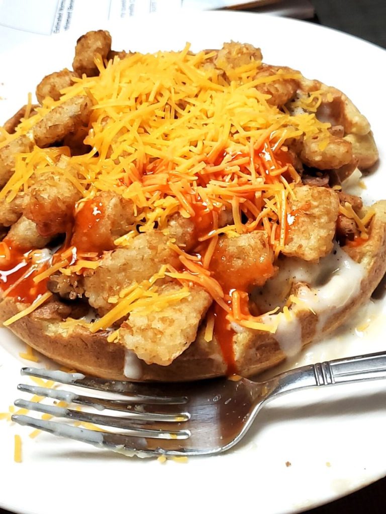 Waffle topped with white gravy, tater tots, shredded cheddar cheese on a white dinner plate with silver fork in front. 