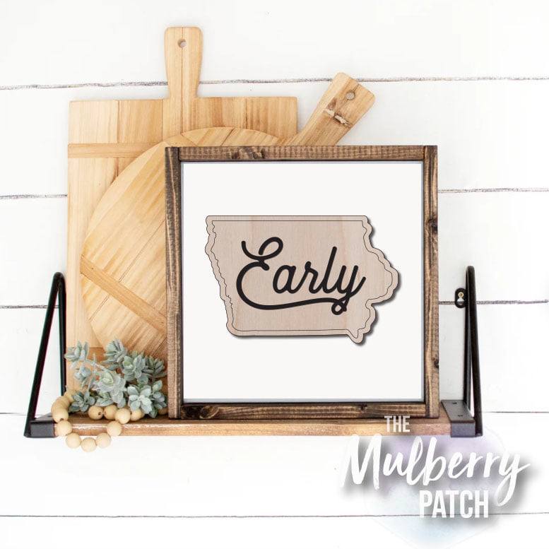 Two wooden cutting boards behind a framed Iowa cutout with the word "Early" in the middle, sitting on a wooden shelf with greenery on the left