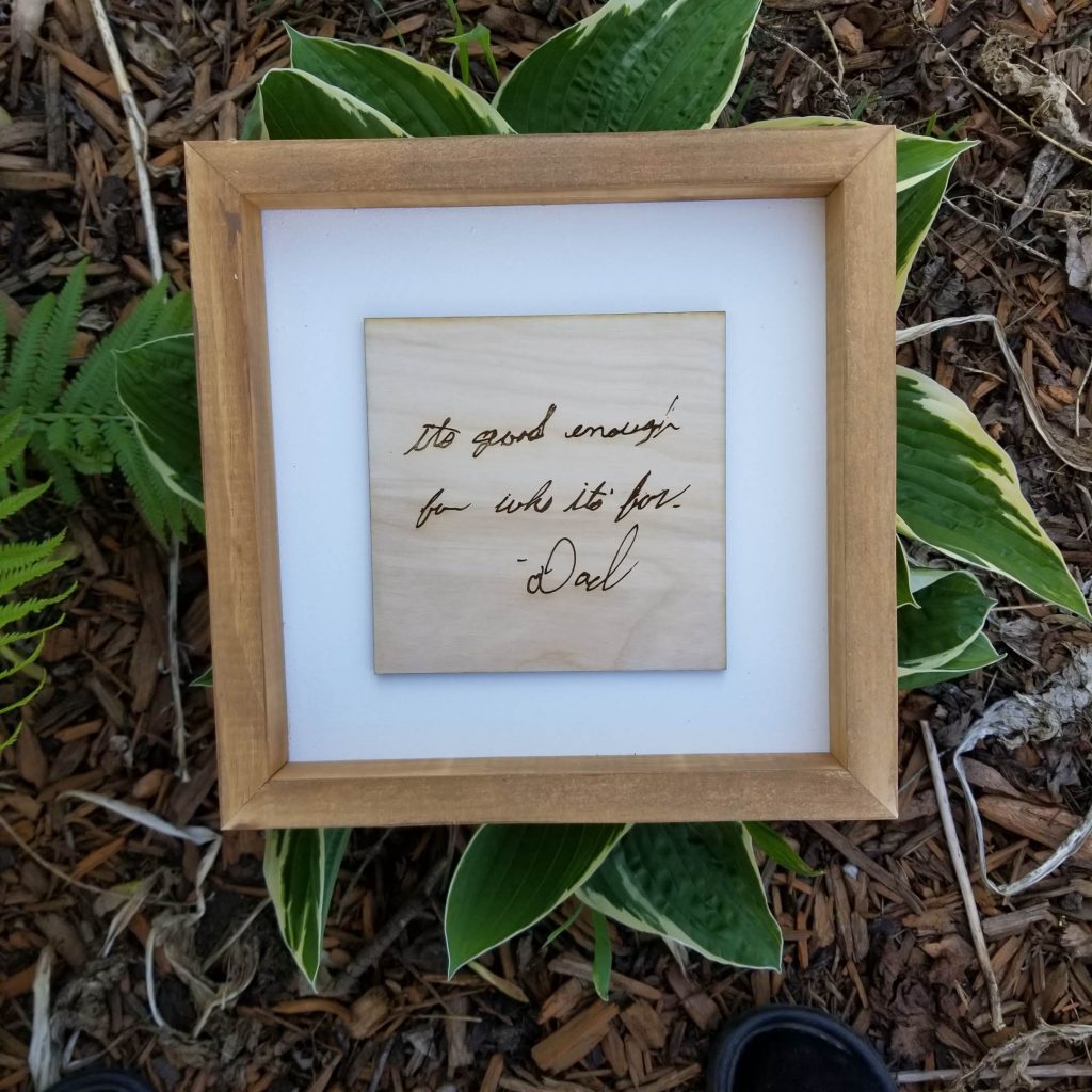 "It's good enough for who it's for." -Dad  laser engraved on a wood, white matting in a wooden frame sitting on a green and white hosta. 