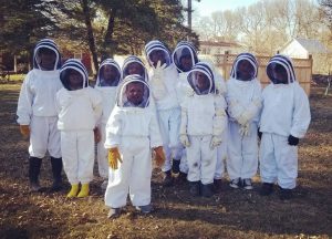 Ten children beekeepers with white suits with black veils with a wooden lattice fence and trees in the background. 
