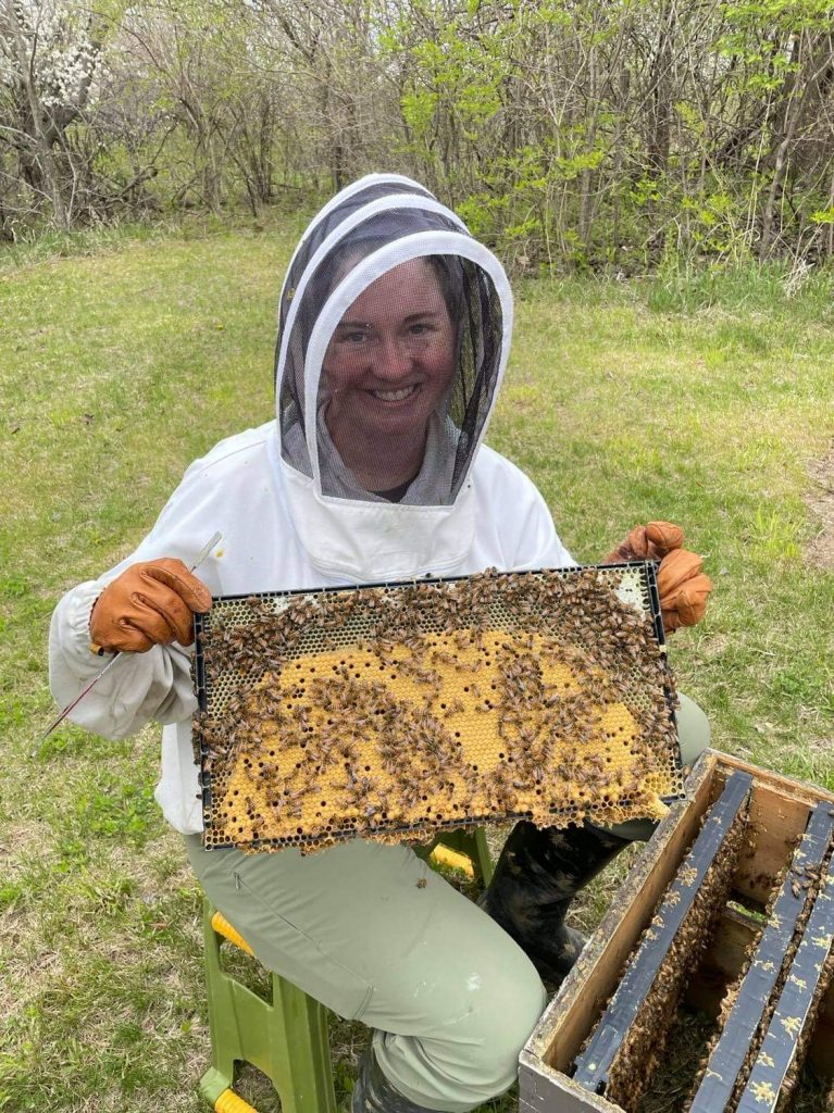Woman holding a frame of yellow honey bee brood in a white and black beekeeping suit with green grass and wooded area behind