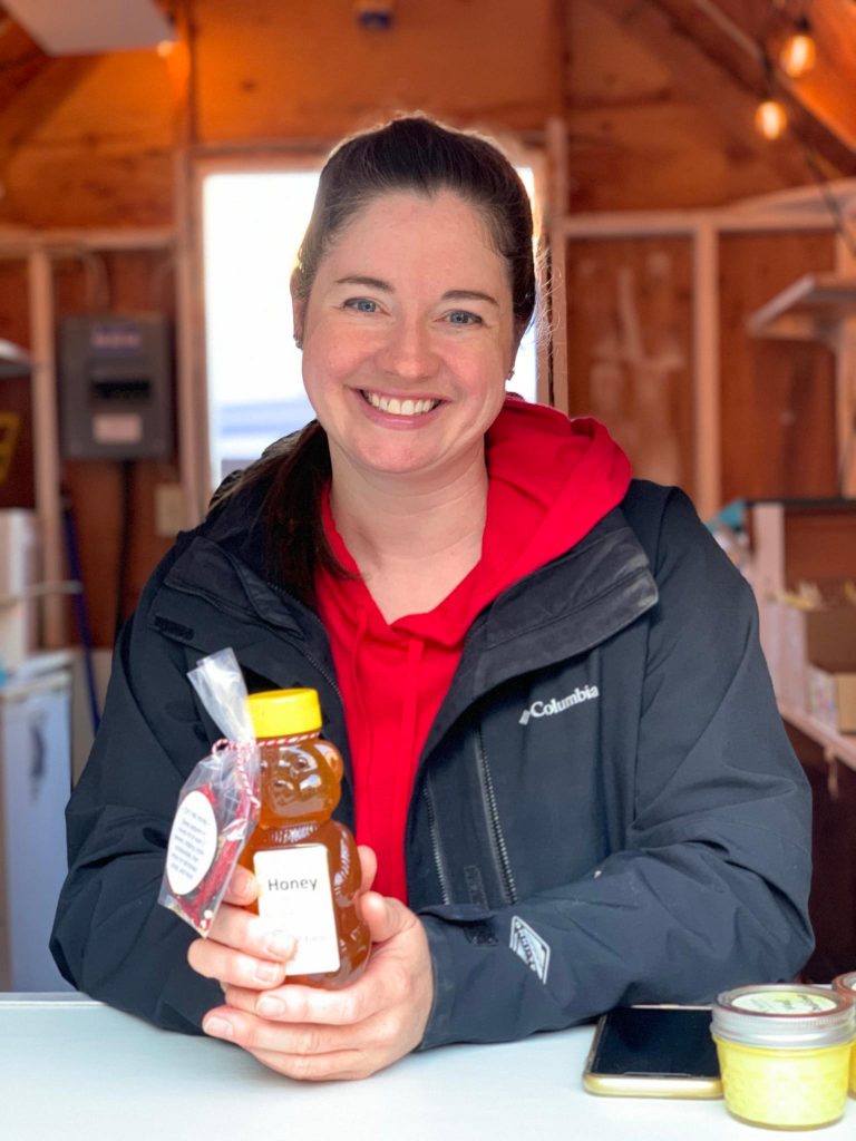 Woman with brown hair smiling holding a 12 oz honey bear in a black jacket with red sweatshirt in a wooden barn