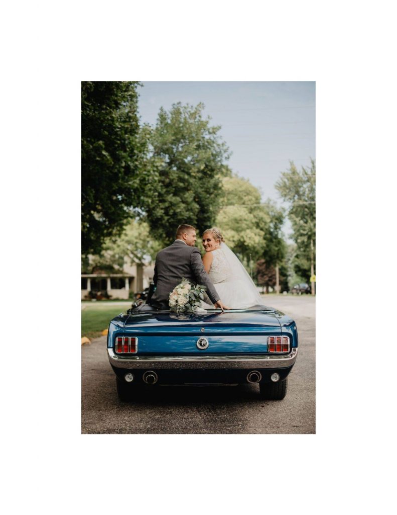 Back end of a blue mustang with groom sitting on the back seat of the car on the left and bride on the right with a bouquet of flowers behind them