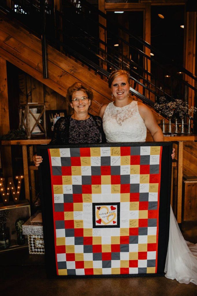woman with brown hair and glasses on the left, bride on the right holding a white, gray, yellow, red quilt in a black frame