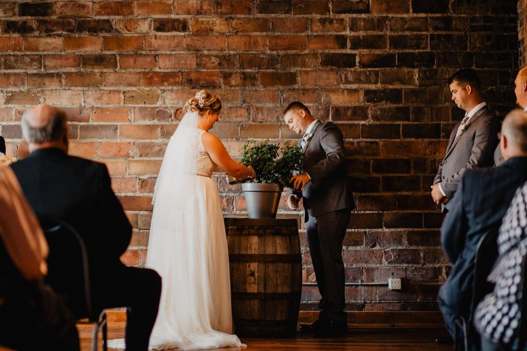 bride on left, groom on right with a plant in a silver pot on a barrel in front of a brick wall.