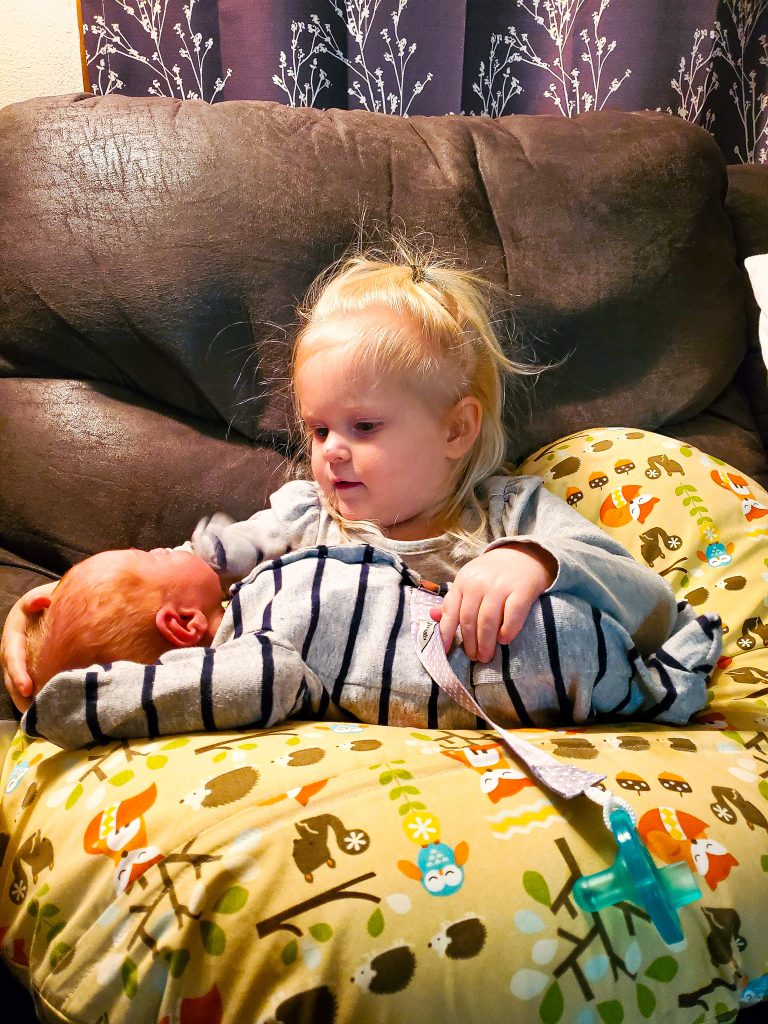 Blonde girl holding a newborn baby in gray striped zip up sleeper on top of an animal print Boppy Pillow sitting on a gray couch in front of gray and white curtains. 