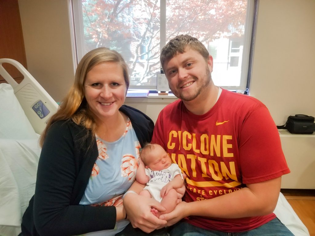 Woman wearing a blue shirt with coral flowers and black sweater on the left with a man wearing a red "Cyclone Nation" shirt with yellow writing holding a newborn baby in front of a window. 