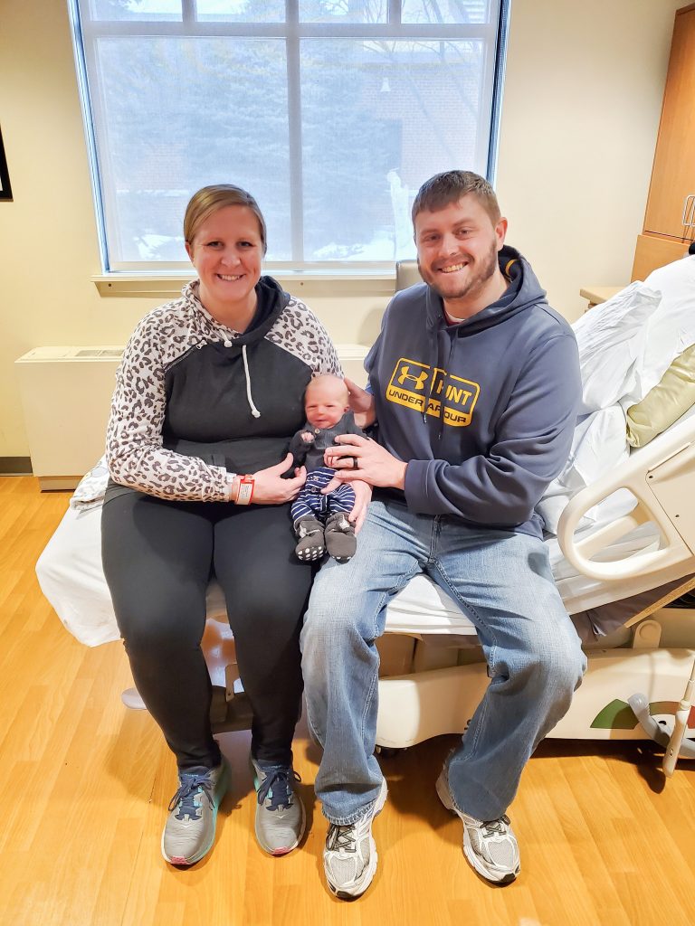 Woman on the left wearing black yoga pants and gray leopard print sweatshirt sitting next to a man on the right wearing a gray under armour sweatshirt with yellow writing holding a newborn baby sitting on a hospital bed in front of a window