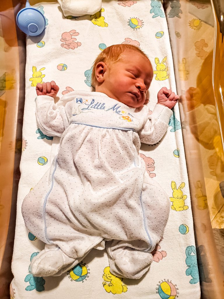 Newborn baby in hospital basinet in a white and blue outfit.