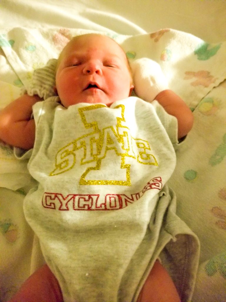 Newborn baby in hospital basinet with Iowa State Cyclones glitter onesie. 