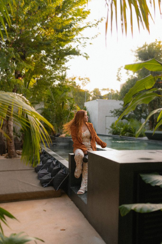 Woman with long orange hair, looking to the right of the picture surrounded by tropical trees, sitting my a pool
