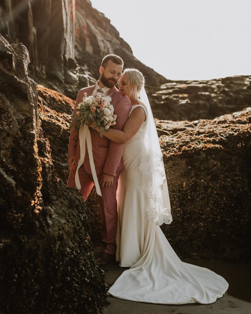 Groom on the left wearing a pink suit with bride with left arm and bouquet with white ribbon on the right wearing a white dress in front of mountains and a sunset. 