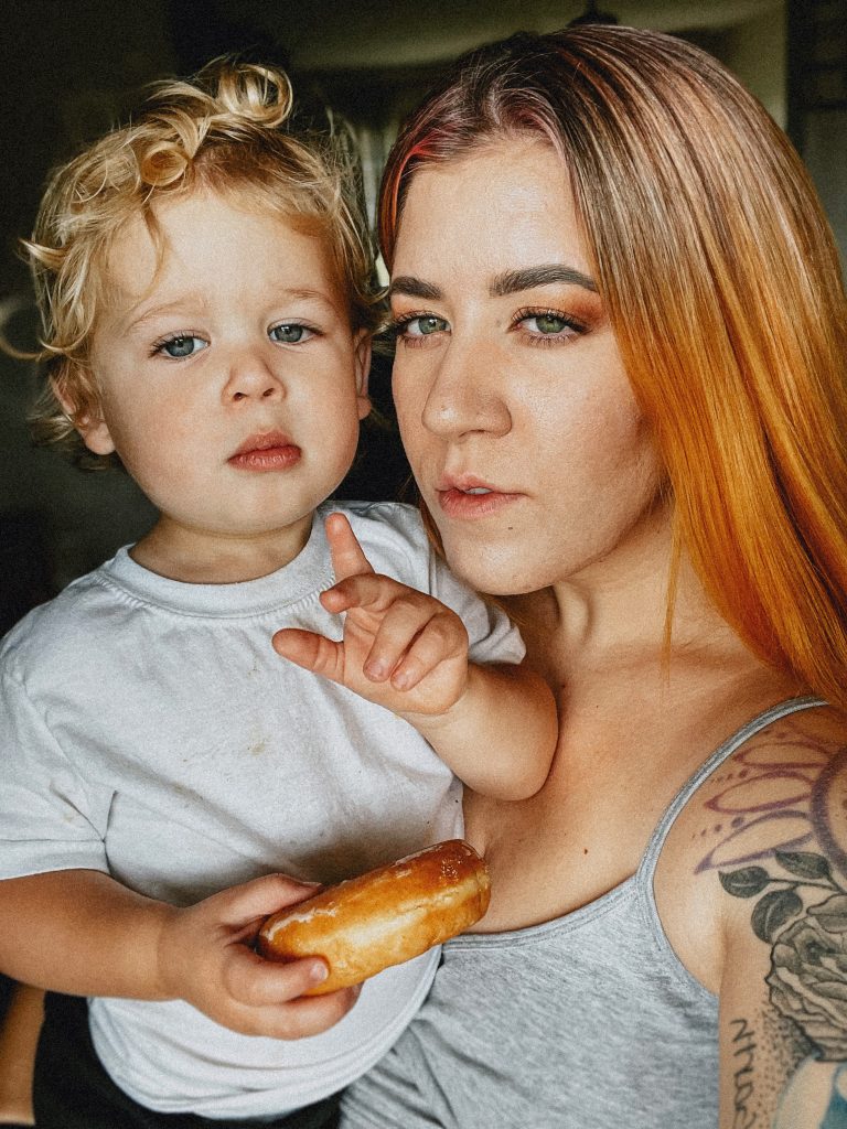 Blonde, curly haired boy wearing a white shirt, holding a donut on the left being held by a woman with orange hair wearing a gray spaghetti strap tank top