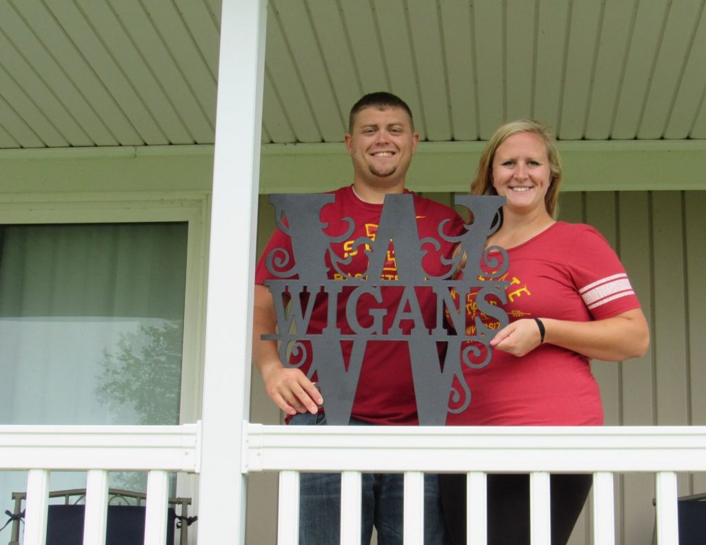 Man on the left wearing an Iowa State University shirt in red and woman on the right wearing an Iowa State University shirt in red both holding a "W" "Wigans" sign behind a white banister and tan background. 