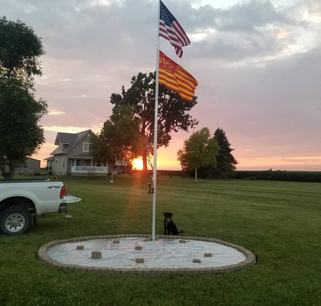 The tail end of a white pickup on the left, next to a circle flag pole area with an Iowa State University and US Flag in front of trees, sunset and tan house. 