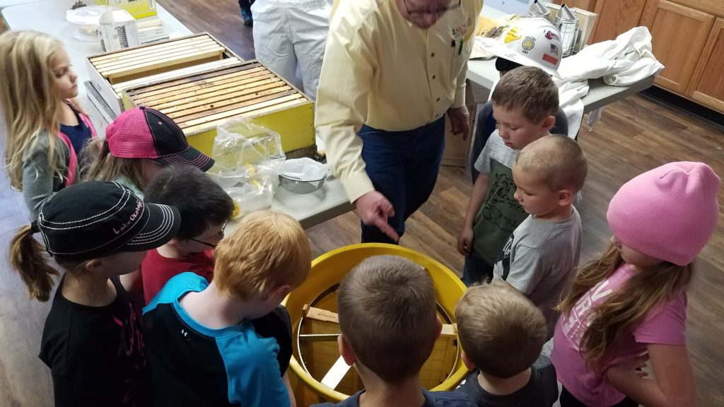 Group of students surrounding an older man wearing a yellow shirt showing the inside of a yellow honey extractor with a hive box and frames in the background. 