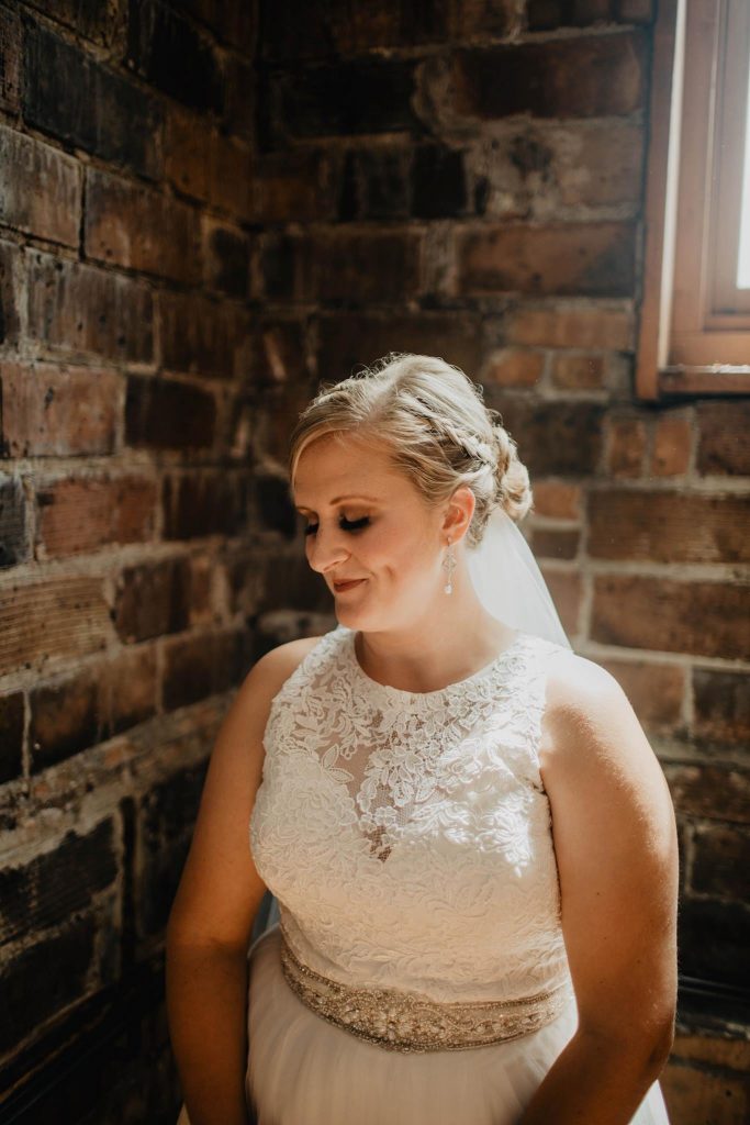 Bride in white lace dress with rhinestone and pearl belt and veil in front of a brick wall