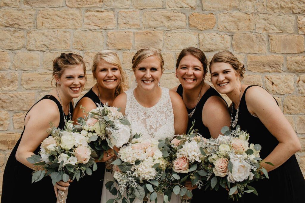 Two women on the left wearing black dresses holding bouquets, bride wearing a white lace dress in the middle and two women on the right wearing black dresses holding bouquets. 