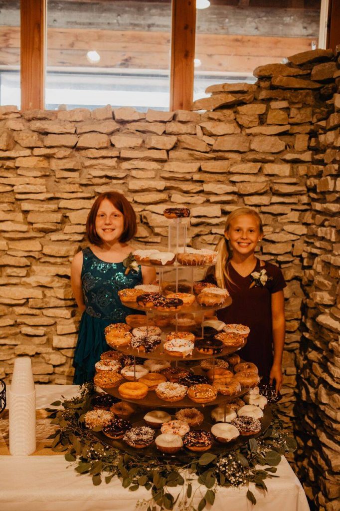Girl in a teal sequin dress on the left, tower of donuts in the middle and girl in a maroon dress on the right in front of a hand chiseled rock wall