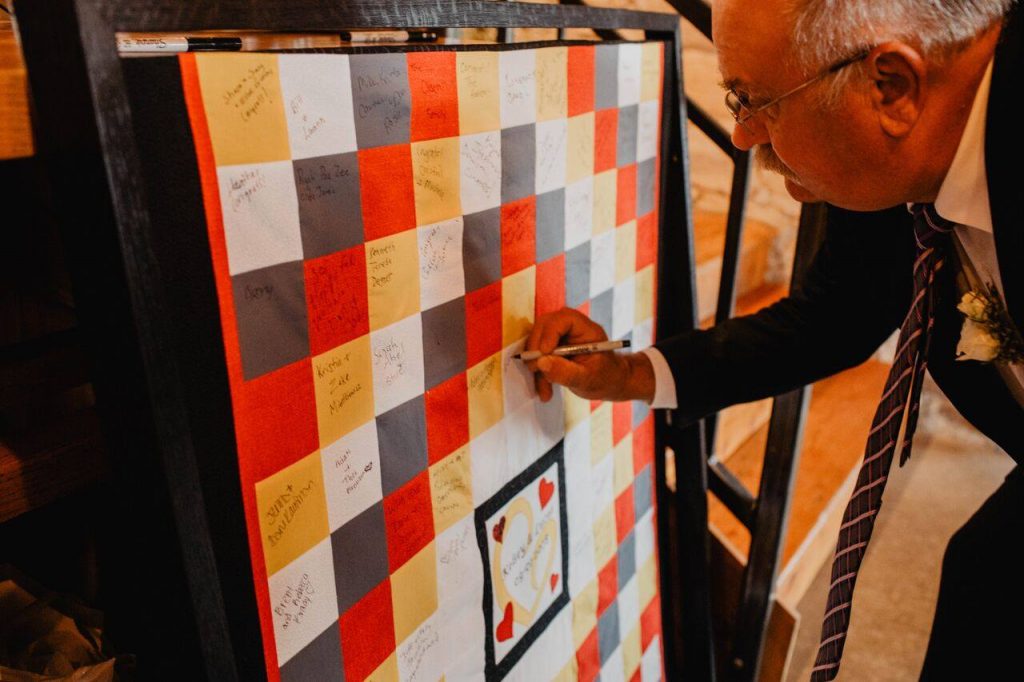 Man with glasses and gray hair signing a yellow, white, gray and red block quilt. 