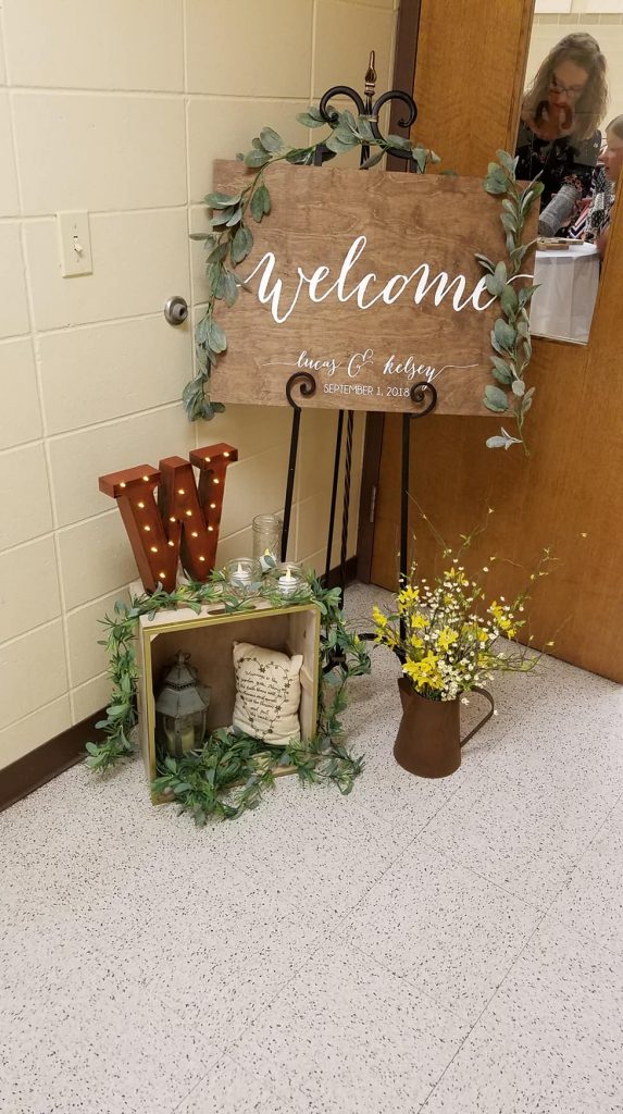 Wooden sign with the word "welcome" in white with various greenery and yellow floral decorations and metal light up "W"