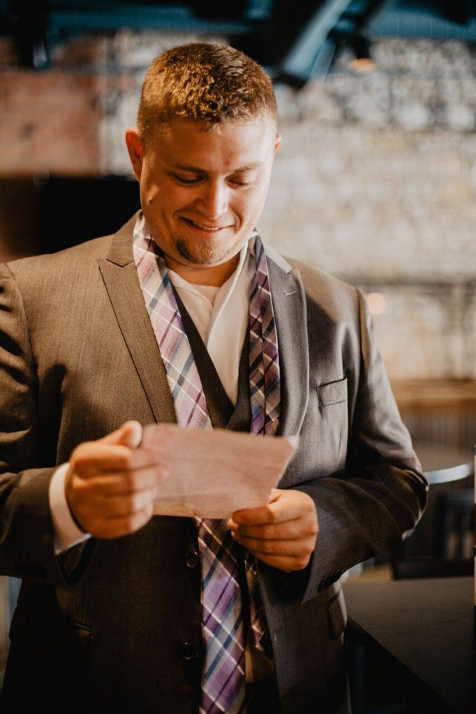 Man with a gray checkered print untied tie around neck in a gray suit reading a note and smiling 