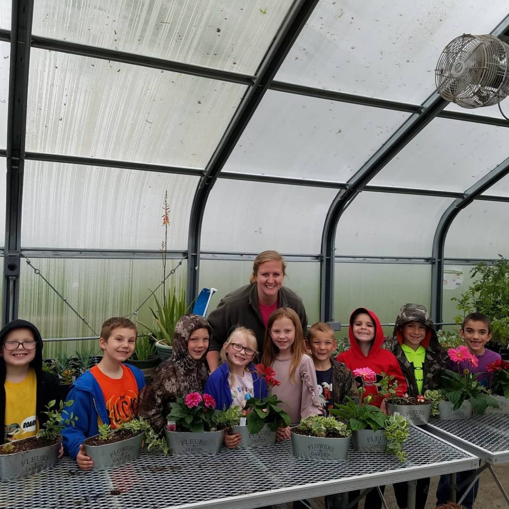 Young students in front with floral arrangements in buckets with teacher in the background. 