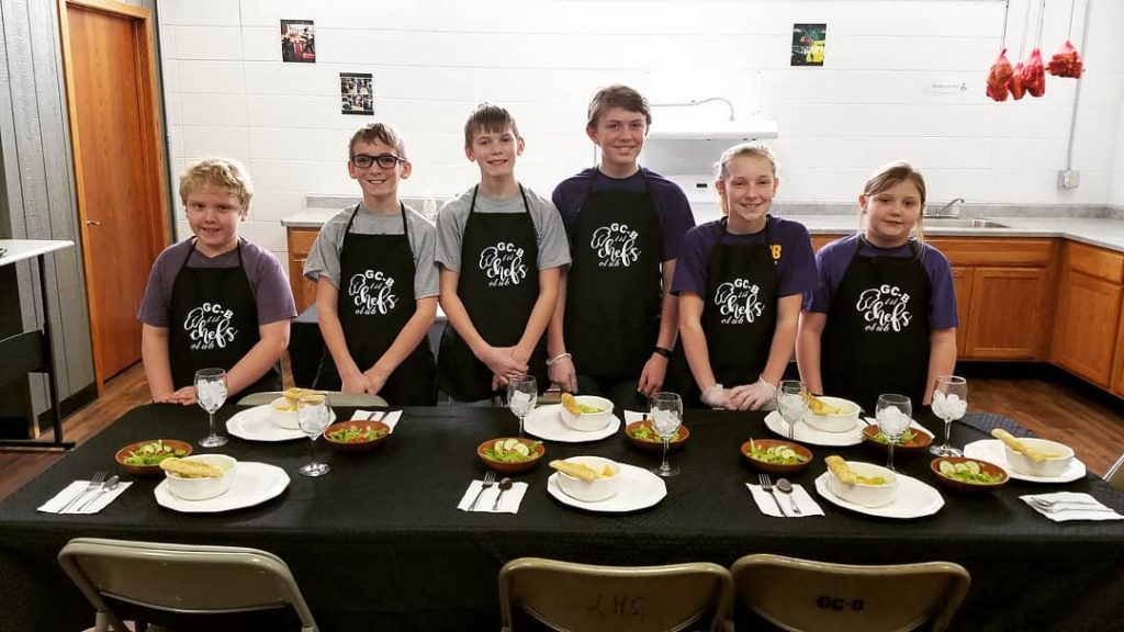 Four teen aged children wearing black aprons in front of a table setting with black table cloth, three boys and one girl