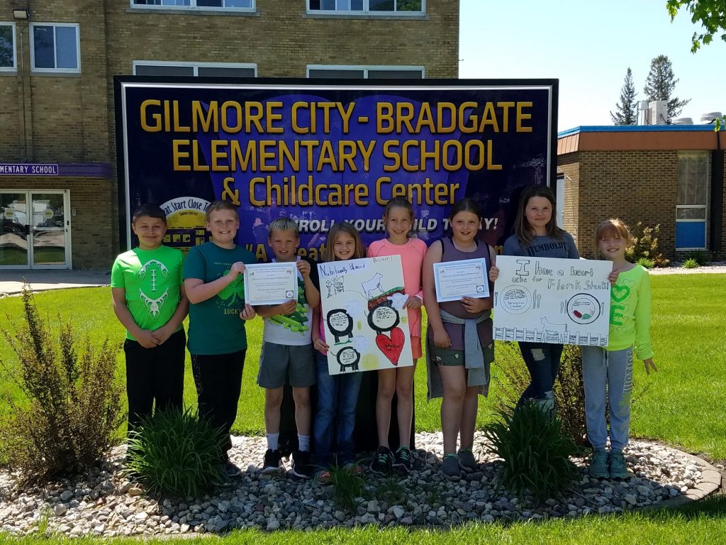 Eight students standing in front of Gilmore City-Bradgate Elementary School sign in front of a tan brick building and grass. 