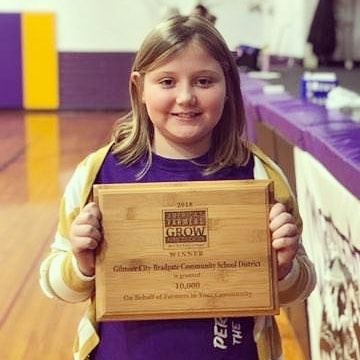 Young girl holding a wooden plaque in front of purple, white, black and yellow mats behind her in the plaque. 