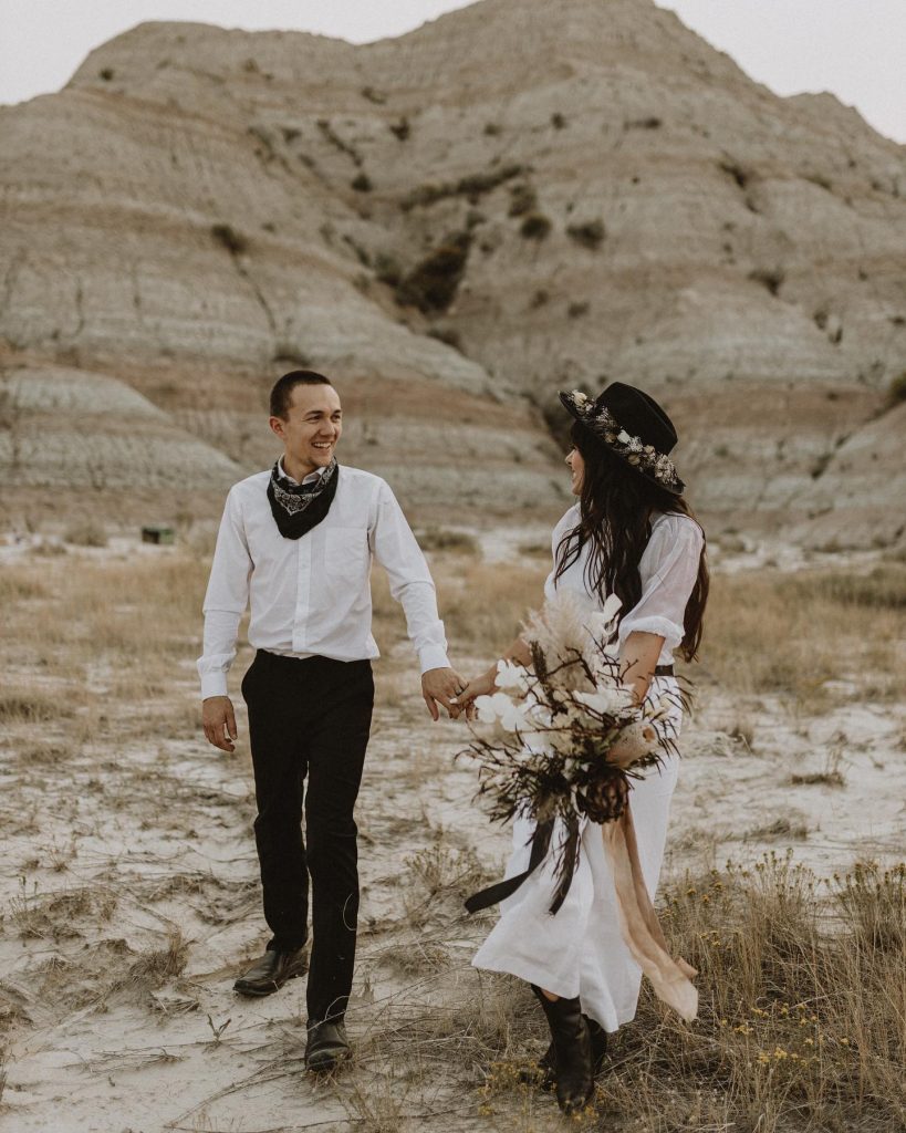 Groom on the left wearing white long sleeve shirt, bandana, black pants with a bride on the right wearing a black wide brim hat and boho inspired bouquet in front of a sandstone mountain. 