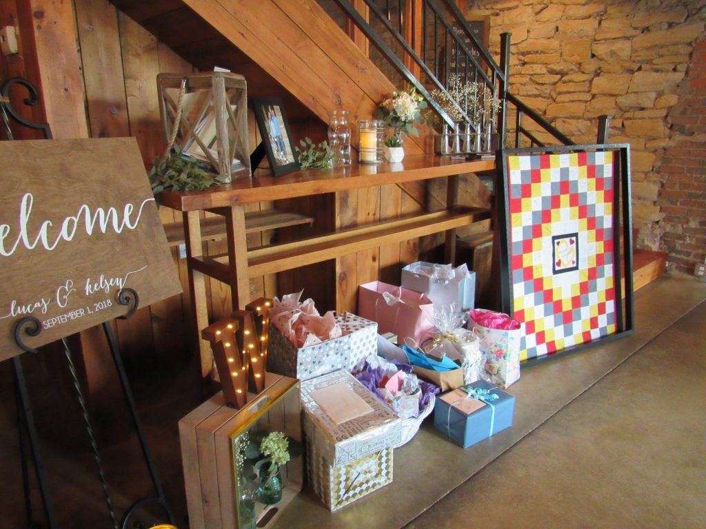 "welcome sign on the left surrounded by presents with a light up "W" with a tall table with decorations and a gray, red, white, and yellow quilt on the right. 