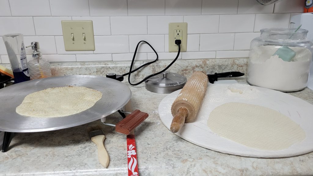 White granite countertop with two round pans of dough with a rolling pin in from of white subway tile wall with a container of flour to the right. 