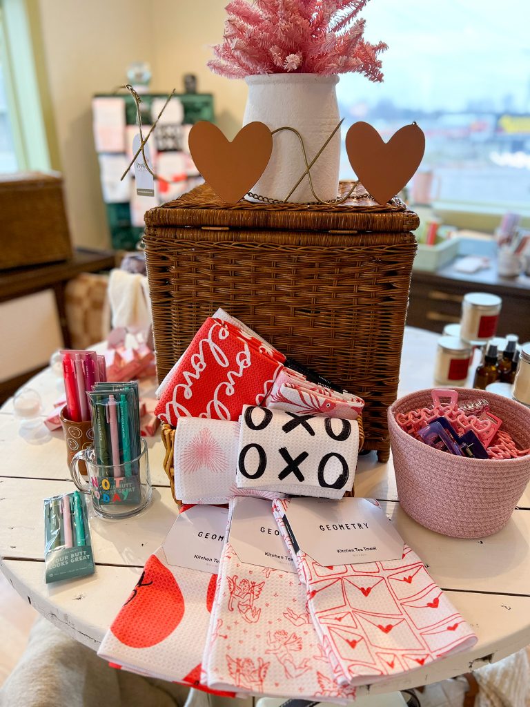 Valentine's Day items sitting on a white table including red heart towels, a pink bowl of hair clips, a brown wicker basket with a white vase with pink faux florals on top. 
