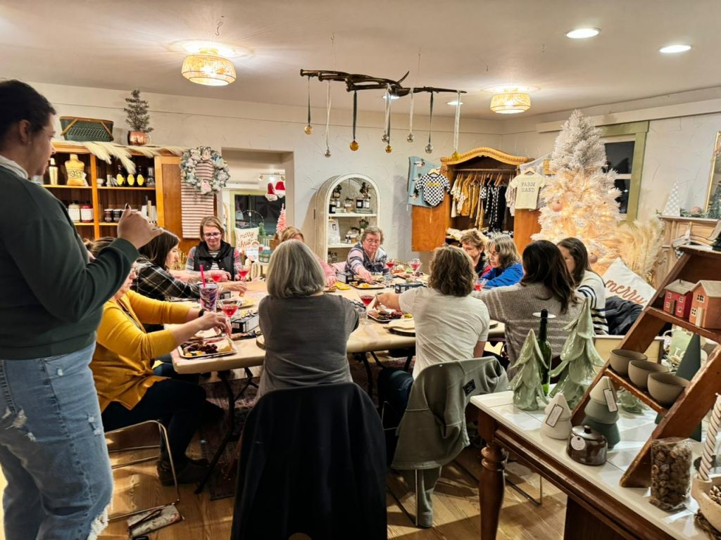 A group of several women sitting around a table smiling enjoying charcuterie boards with Christmas and winter decorations in the background. 
