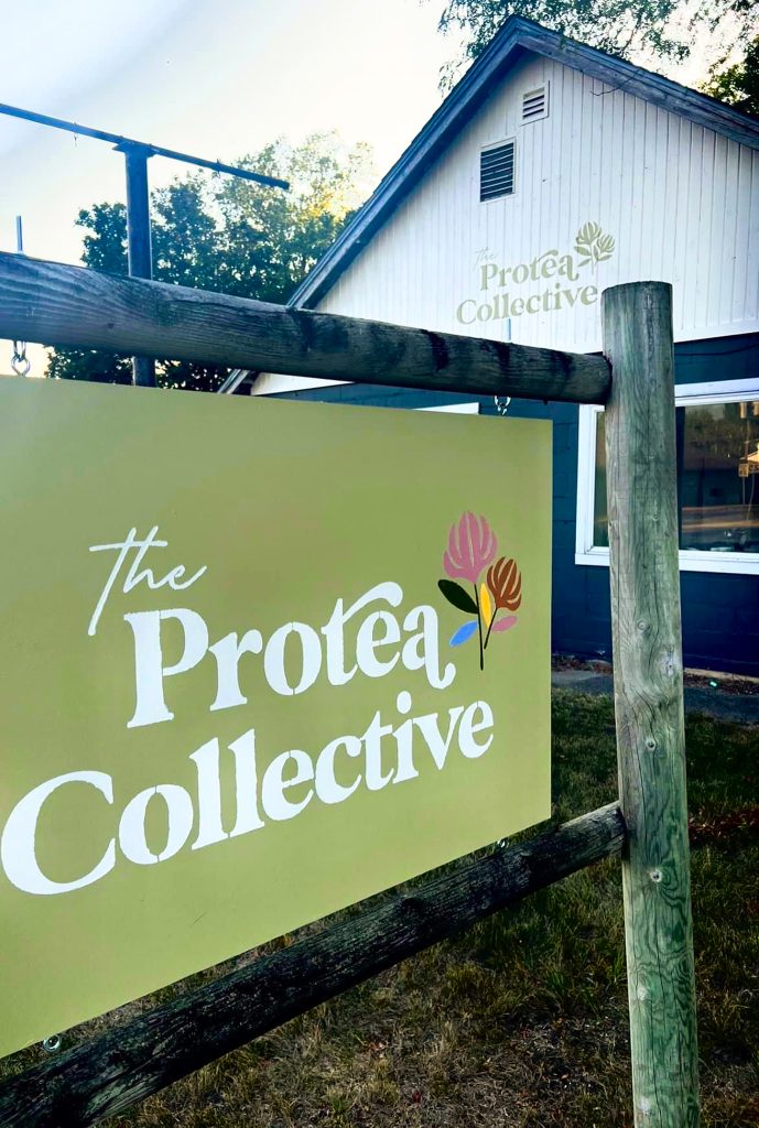 A green sign with the logo containing the words, "the Protea Collective." in front of a gray and white building with the same logo. 